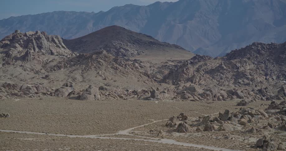 A panoramic view captures a vast arid landscape of rounded rock formations and desert scrub with a towering mountain range visible in the hazy distance