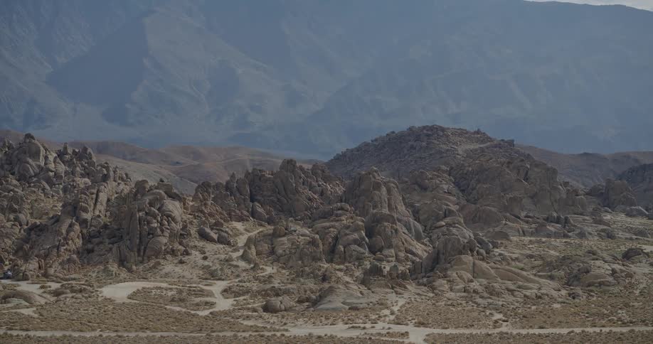 A vast arid landscape features a foreground of rolling rounded rock clusters and flat sparse ground with vehicle tracks set against a background of much larger distant mountains shrouded in a light haze