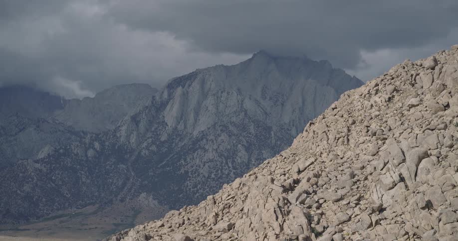 Sunlit rocky slope in the foreground with shadowed mountains under cloudy sky in the background