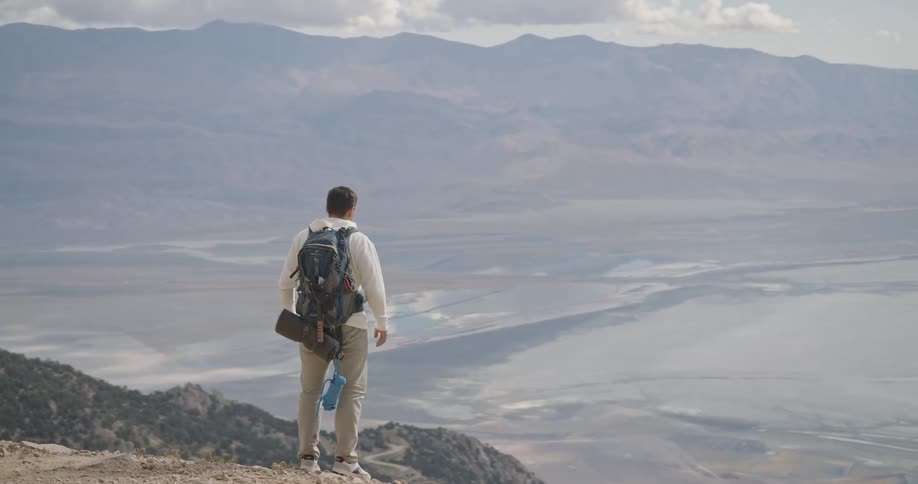 A male hiker wearing a backpack sits on the edge of a mountain cliff overlooking a vast valley and distant hazecovered landscape he rises to his feet and gazes out at the sweeping expansive view of the rugged terrain