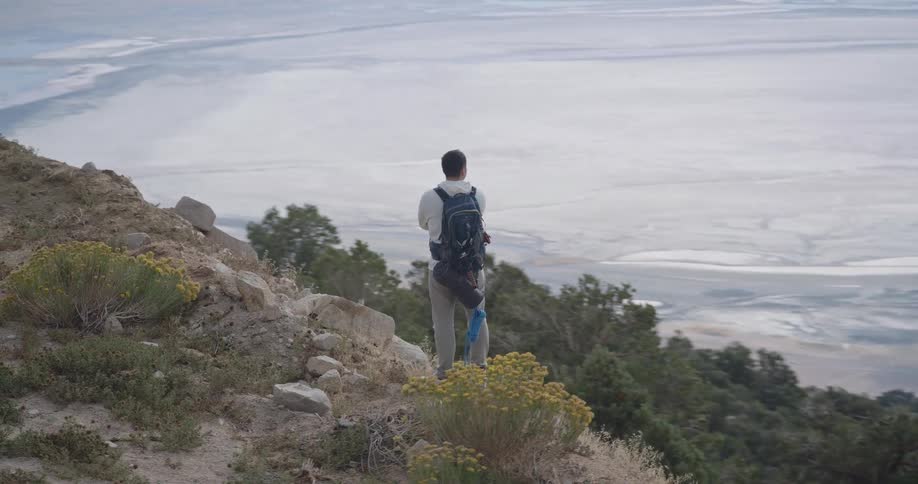 A male hiker stands on a rocky slope above green shrubs gazing contemplatively over a vast pale valley floor far below