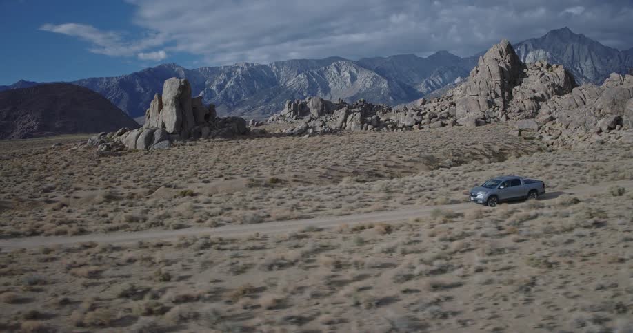 A gray pickup truck drives away from the camera on a dusty dirt track traversing a wide brushcovered desert valley dotted with large rounded rock formations while towering mountain peaks stand against a partly cloudy sky in the background