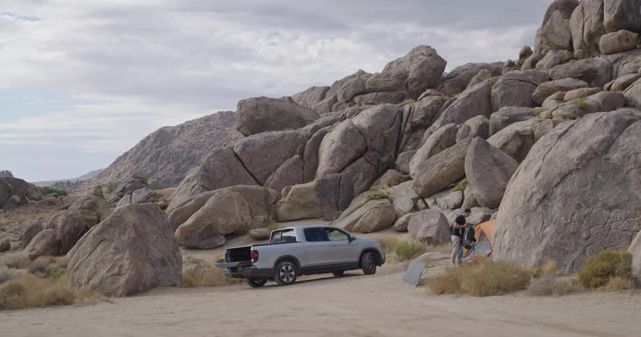 A man stands beside a silver pickup parked among giant rounded boulders in a desert pullout under bright patchy clouds