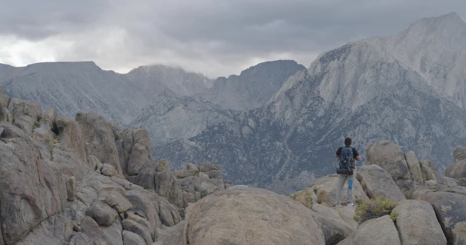 A lone hiker with a blue backpack stands atop rugged boulders facing towering granite mountains beneath a moody overcast sky