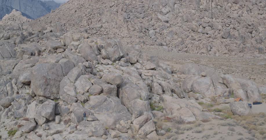 The video is a highangle shot of a male hiker climbing down large rounded boulders in a rugged rocky landscapein the upper background there is a gray vehicle parked beside a small orange tent suggesting a remote camping and hiking spot