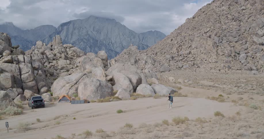 A solo hiker walks towards a campsite with a truck and orange tent toward towering granite formations in a wide sandy basin beneath brooding mountains and clouds
