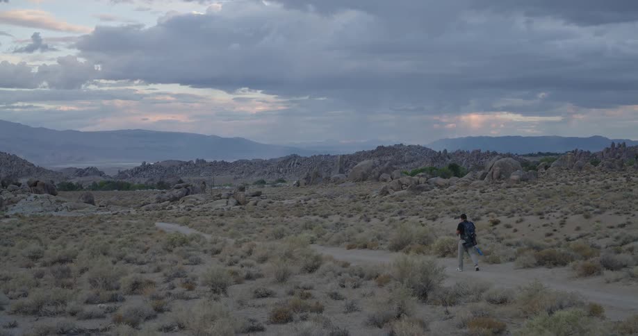 A person with a backpack walks away from the camera along a dirt path through a flat brushcovered field moving toward a formation of scattered rocks and a distant mountain range beneath an expansive cloudy sky