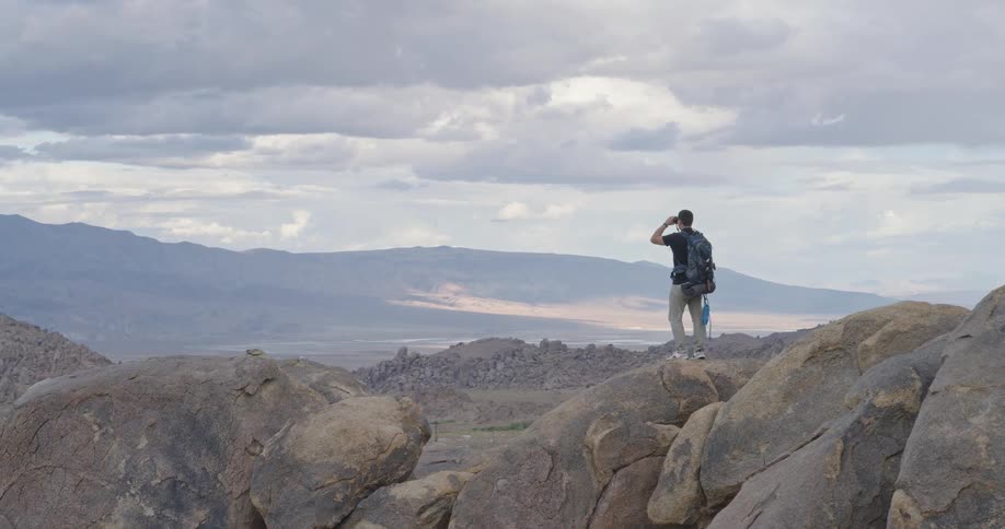 A backpacker stands on a large rocky outcrop looking out at a vast arid landscape with a handheld device possibly a camera or binoculars the camera slowly zooms or tracks out revealing the dramatic scale of the mountain range and valley under a clou
