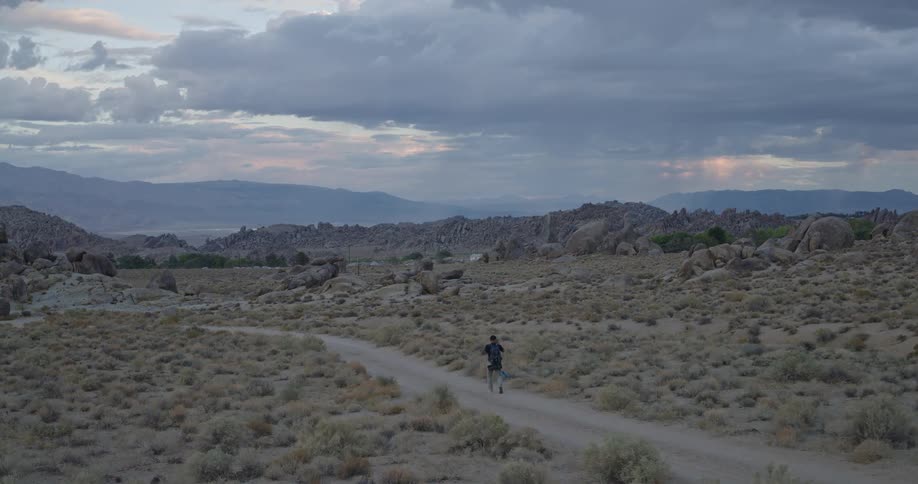 Person walking along trail in desert landscape with dry vegetation and mountains in the background