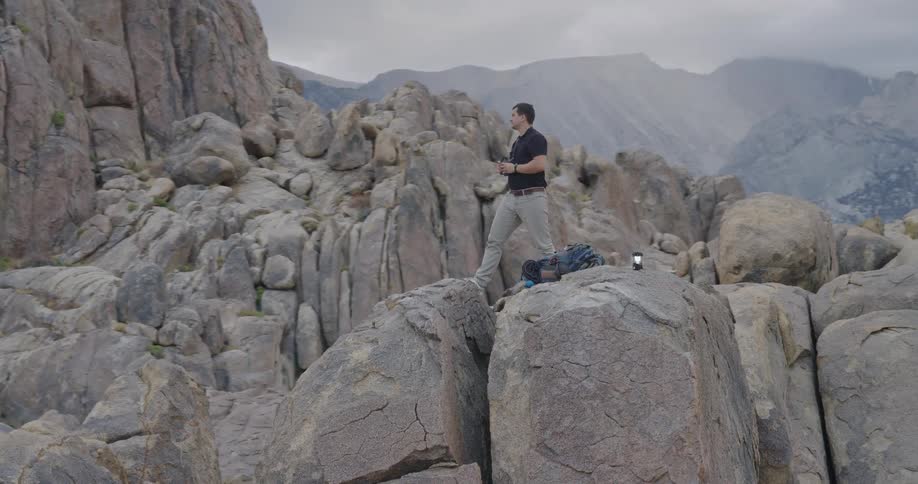 A man stands on a large boulder scanning the rugged mountain range with binoculars appearing focused under a cloudy sky