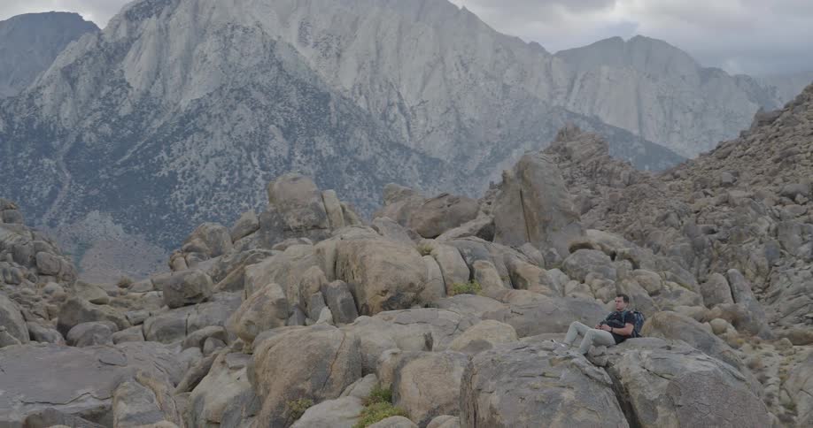 A male hiker lounges on rounded granite boulders using binoculars to sight see beneath towering speckled mountains