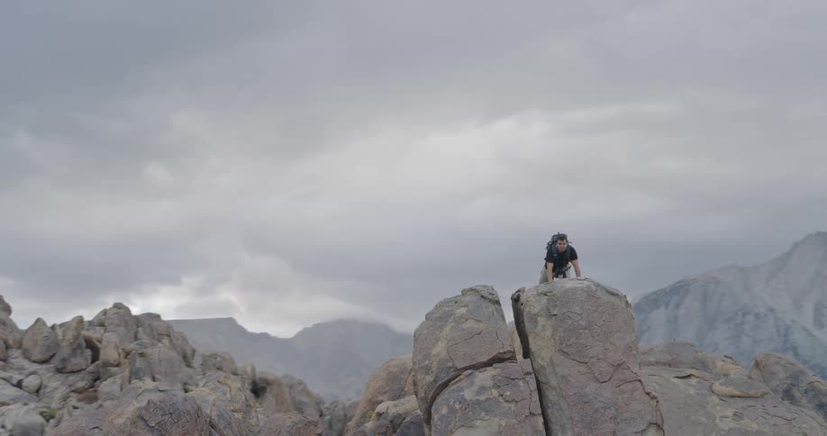 Man lying down in prone position on a high rock looking through binoculars in a region surrounded by mountains outdoors during day time