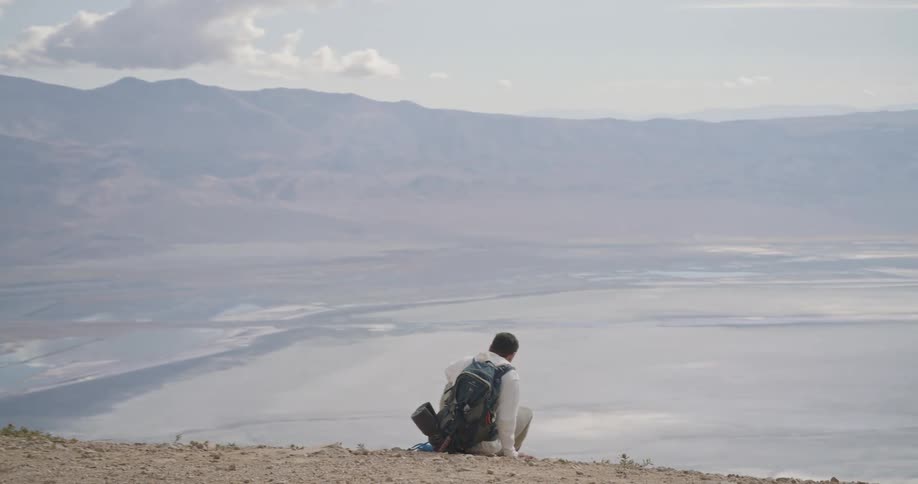 Person sitting on elevated trail overlooking salt flat surrounded by mountains