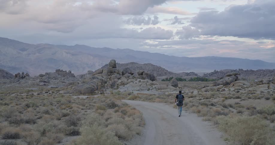 Man walking along trail in dry terrain with rock formations and mountains under partly cloudy sky