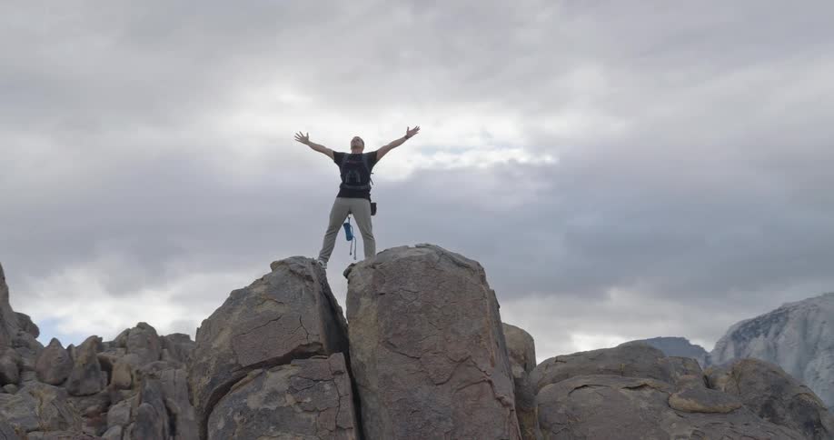 A man stands atop a prominent rock formation in a field of boulders raising both arms outward as the camera pulls back and circles revealing a dramatic mountain environment under a cloudy gray sky