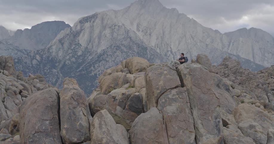 Person with backpack sits on boulders with mountains behind under a cloudy sky