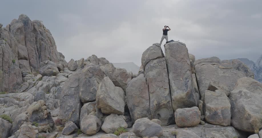 A man standing on a large rock formation looking through binoculars at the dramatic rocky mountain landscape under a cloudy sky
