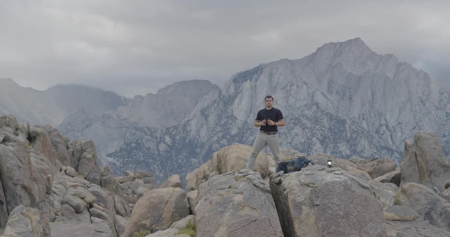 The video shows a male hiker standing on a large rugged boulder in a mountainous rocky landscape he is looking through a pair of binoculars at the distant hazy mountains that form the backdrop