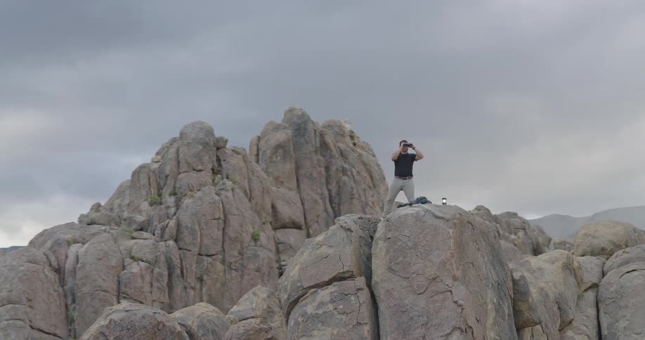 A lone man stands atop a massive boulder using binoculars silhouetted against a gray overcast sky in a rugged rocky landscape