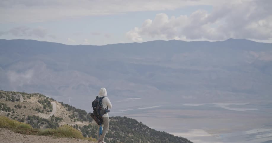 The video shows a male hiker standing on a dirt trail on a steep green mountain slope on an overcast dayhe is taking a photo or video with his phone capturing the vast hazy valley and the distant winding road on the mountainside