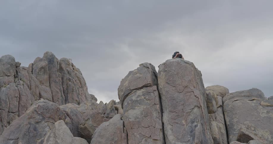 A man lying on a large boulder looking through binoculars at the vast rugged mountain landscape surrounding him