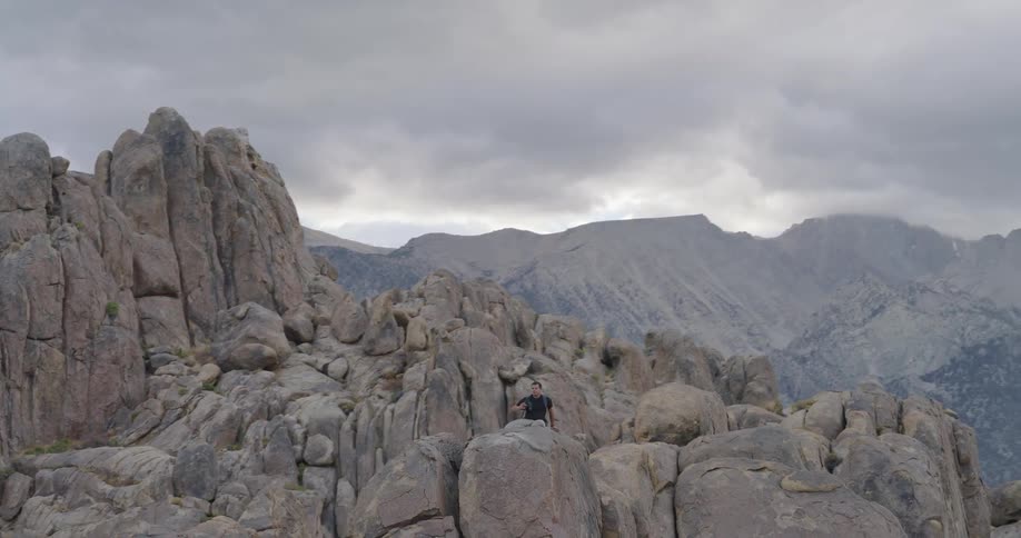 A male hiker walks then sits among rounded desert boulders with rugged mountains behind looking relaxed and thoughtful