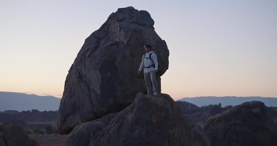 The video shows a male hiker standing atop a large prominent rock formation at dusk or dawn he is looking out at the vast desolate mountain landscape with a hazy grayblue backdrop of distant peaks and scattered boulders