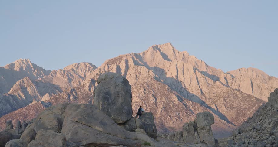 A lone hiker stands on top of a large rounded boulder with their back to the camera looking out over a vast valley and the towering arid peaks of a mountain range the scene is captured during the golden hour bathing the rugged rocky landscape in war