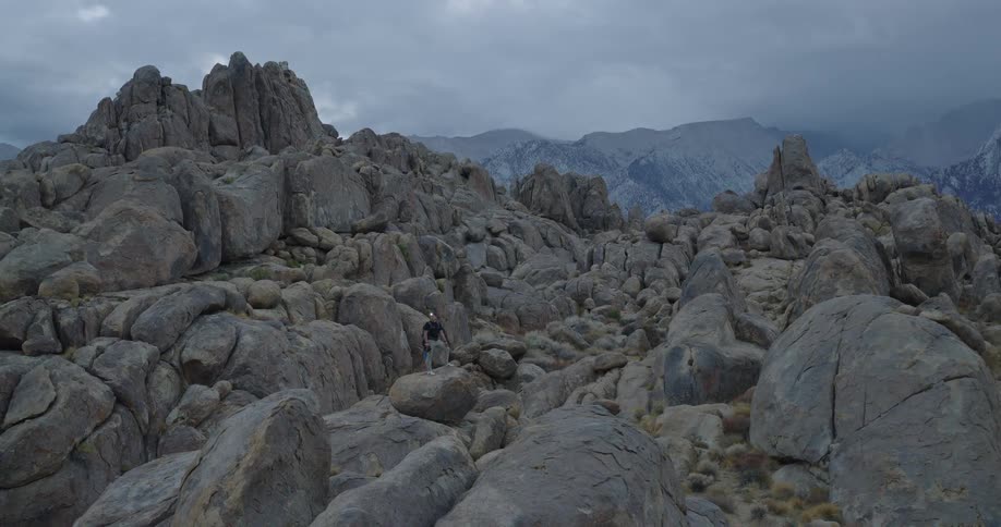 A man stands on a large rock in the foreground then steps down and proceeds to walk through a dense field of large rounded boulders and uneven ground with a large mountain range visible in the far distance under a heavily clouded sky