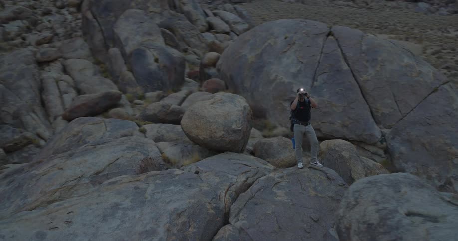 Man with backpack using binoculars on rock in desert area with sparse vegetation