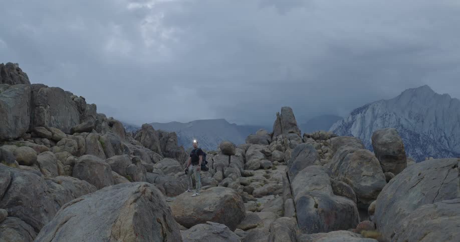 A man with a headlamp on stands balanced on a large rounded rock in the middle of a dense field of similar boulders looking toward the camera as the scene reveals a large dark mountain in the far distance under a heavily clouded gray sky