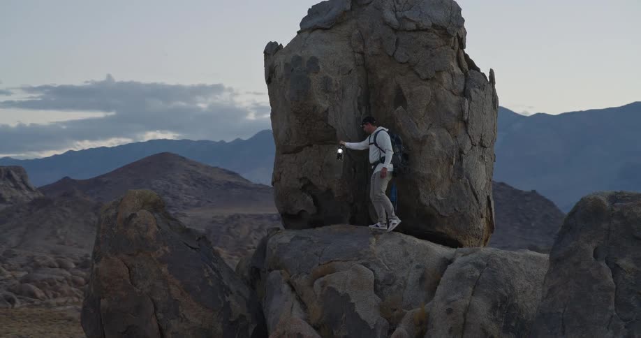A man with a backpack and cap carefully steps along a high rocky outcrop at dusk holding up a small flashlight toward a towering boulder