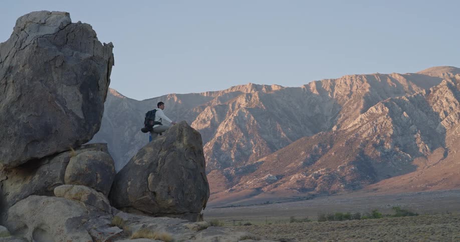 The man is leaning on a large boulder by the mountains