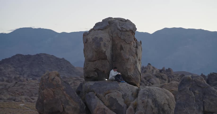 A man sits at the top of a rocky mountain in the wilderness at sunset during a hike