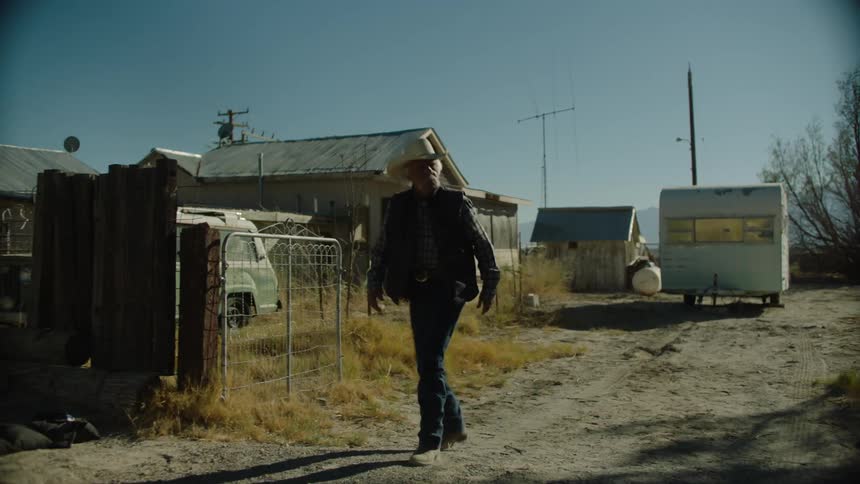 A man with a cowboy hat is walking down a dirt road to a house
