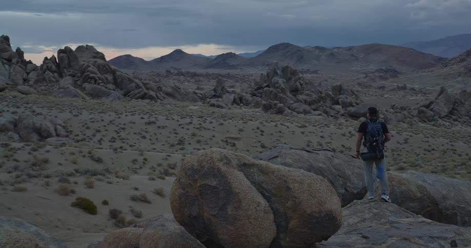 The video is a wide establishing shot of a rugged rocky and desolate mountain landscape under an overcast sky a hiker with a backpack is visible traversing the large rounded boulders in the foreground as the camera slowly pulls out highligh