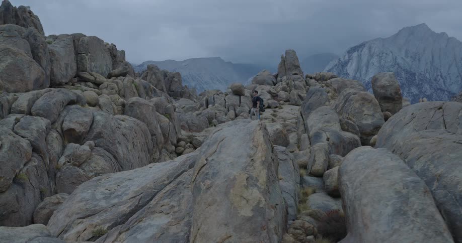 The video is an extreme long shot showing a hiker traversing a dense field of large rounded gray boulders under a heavily overcast skythe enormous scale of the jagged mountain backdrop emphasizes the challenging and desolate nature of the