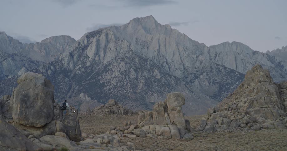The video is an extreme long shot showing a hiker ascending a large rounded boulder in a vast arid and rocky landscapethe background is dominated by a majestic towering mountain peak that is illuminated by soft natural light likely from t