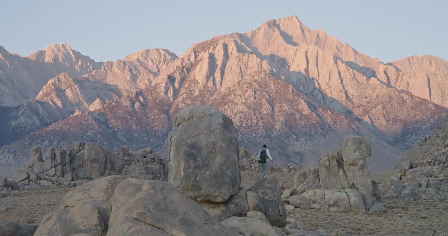 The video is an extreme long shot of a solitary hiker standing on a small rock formation in a vast arid landscape the background is dominated by a majestic sunlit mountain range of jagged peaks under a clear sky
