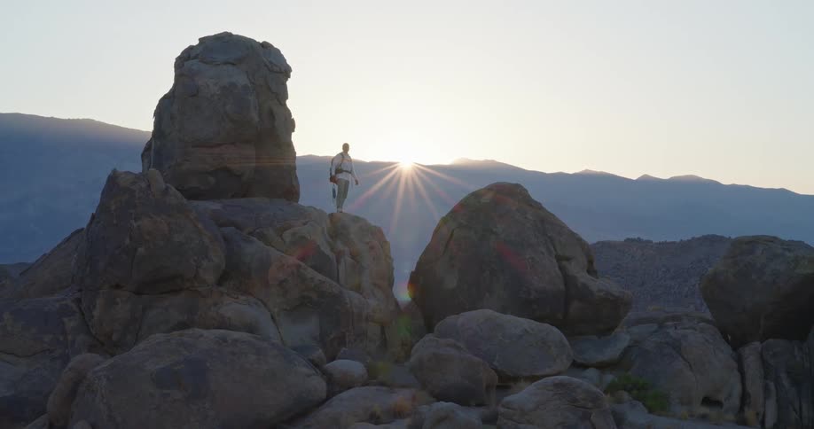 Person standing on elevated rock with sun rays behind among mountains