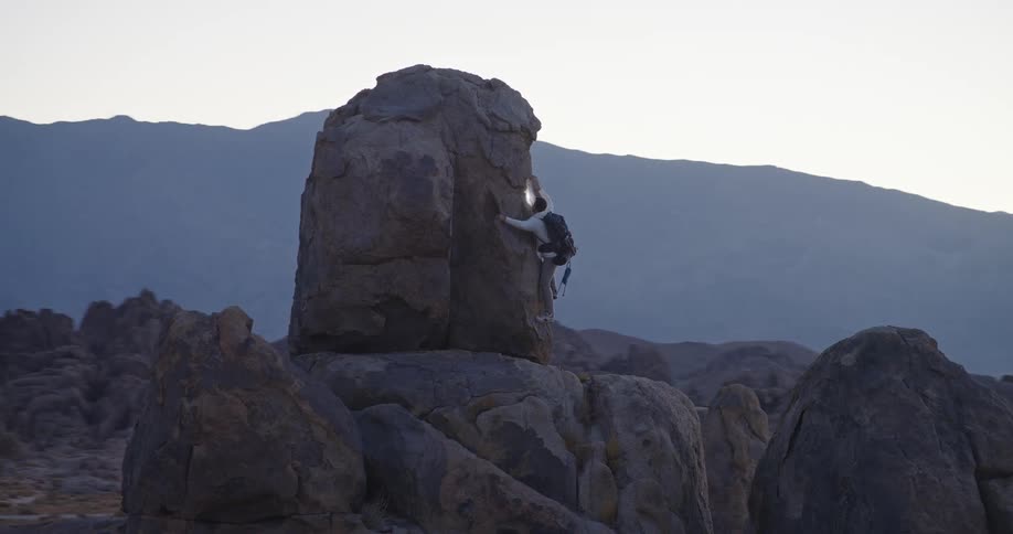 Person climbing sloped rock surrounded by mountains under soft light