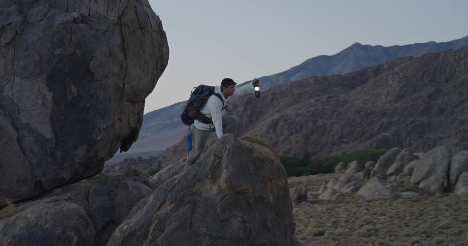 Man with backpack and flashlight on rock in mountainous landscape at dusk