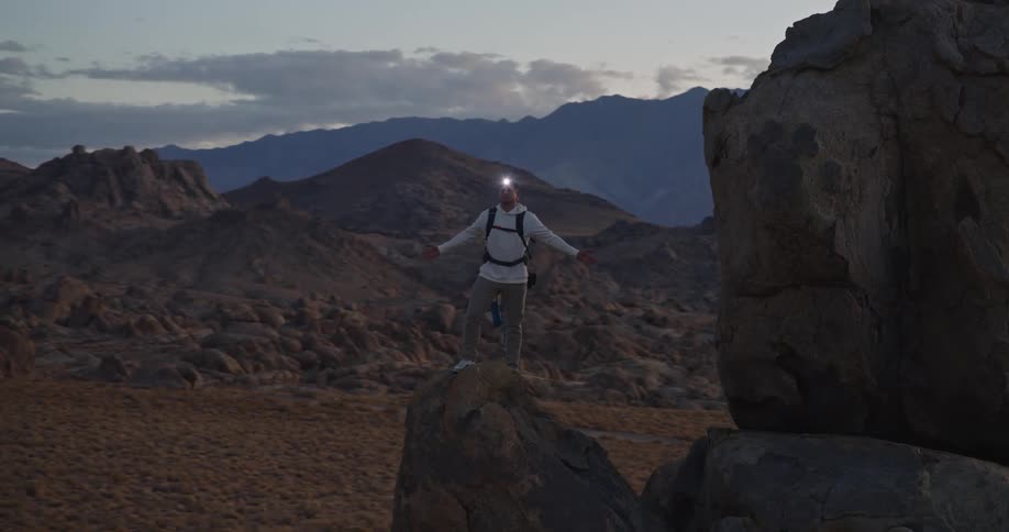 A man wearing a headlamp with the light turned on stands on a narrow rock formation with arms outstretched looking out over a wide dark landscape of hills and distant mountains under a dusky cloudy sky