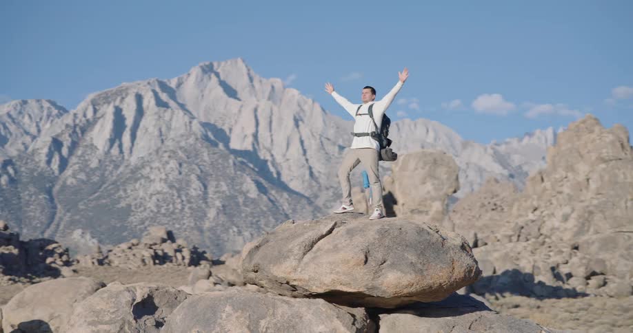 Man with backpack standing on rock formation arms raised in triumph before mountains under blue sky