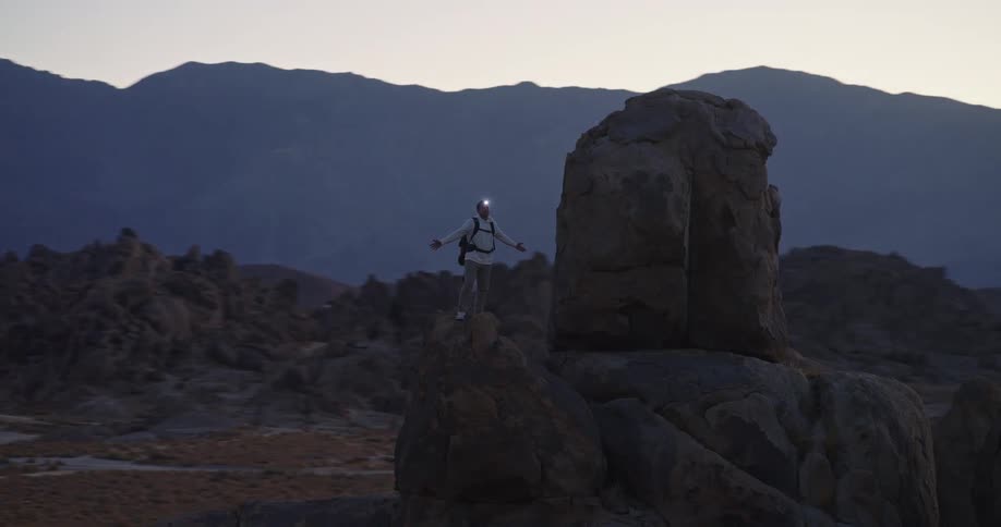 At dusk a male hiker wearing a headlamp stands with arms out on a high boulder above shadowed hills appearing triumphant