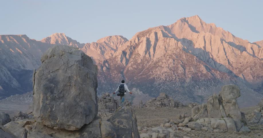 A hiker with a gray backpack stands on a rock outcrop at sunrise gazing at sunlit granite peaks glowing warm against a pale sky