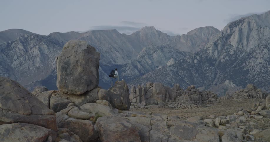 A hiker stands on a large craggy boulder overlooking a vast dramatic landscape of rocky terrain and steep mountains under an overcast sky the overall scene captures the rugged isolation and grandeur of an alpine or desert mountain range