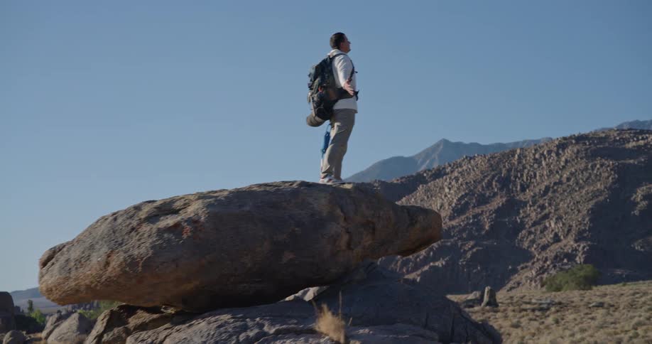 A man wearing a backpack stands on a large rounded rock in the foreground looking out over a wide sunlit landscape of lower hills and distant towering mountains beneath a clear blue sky
