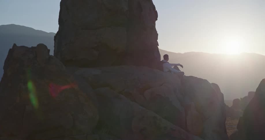 Person seated on rock with legs extended observing horizon at sunset in mountainous area