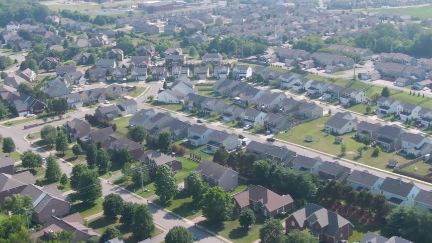 An aerial view shows a leafy suburban neighborhood with rows of houses and curving streets on a bright day